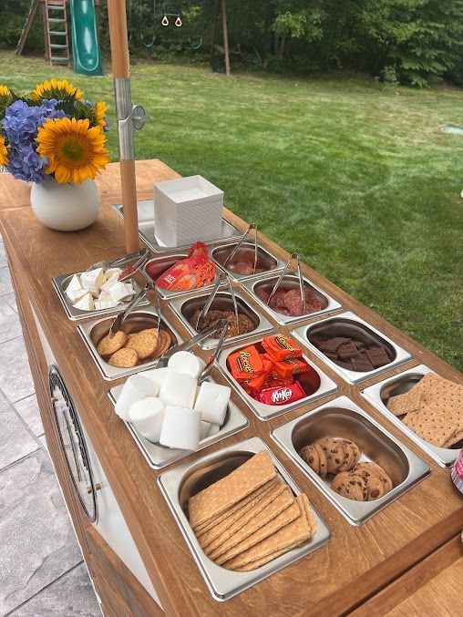 Outdoor wooden dessert table with assorted pastries, fruit, marshmallows, and a vase of sunflowers outdoors