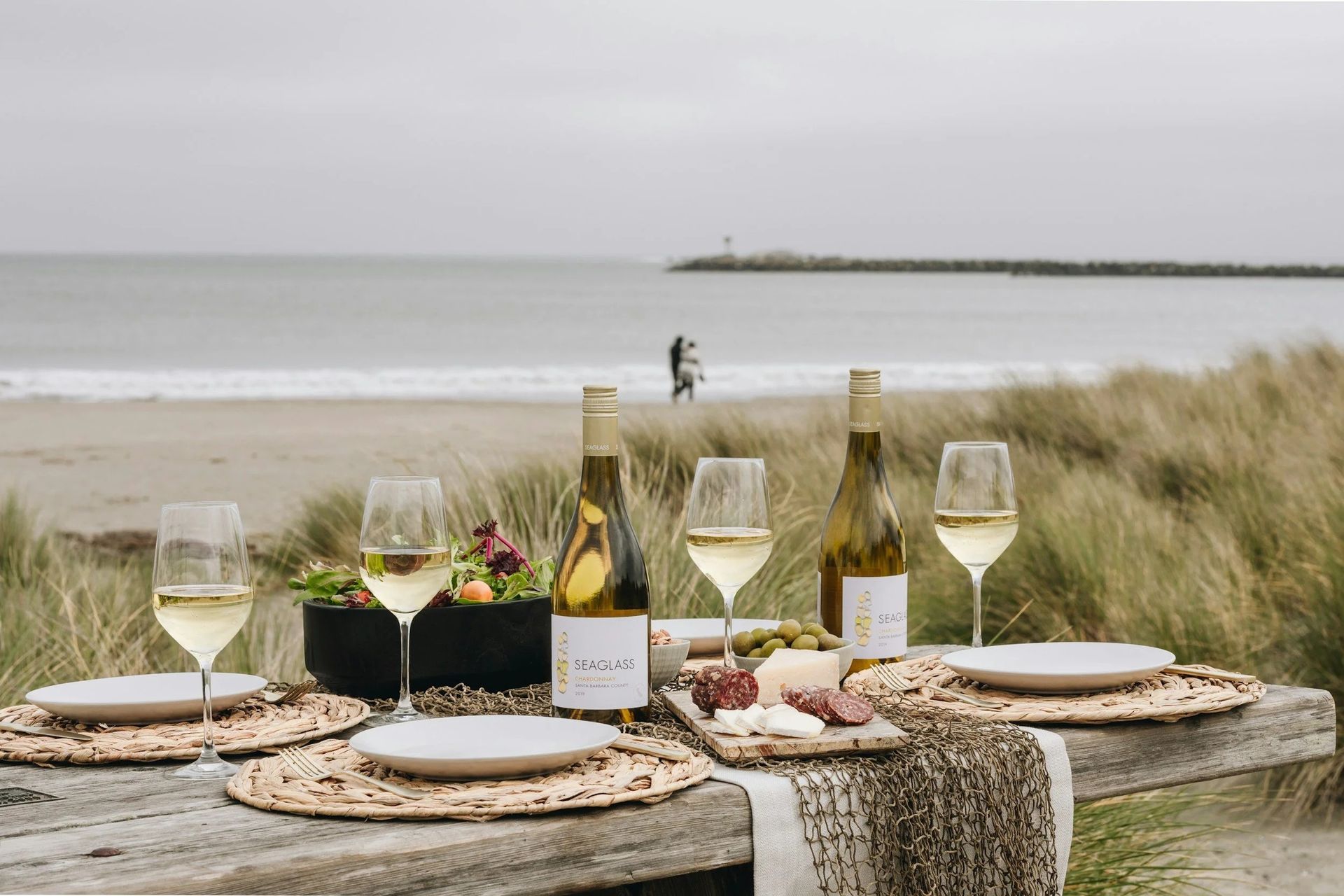 Seaside dining table set with wine glasses, plates, and flowers beside a calm beach