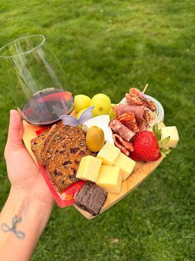 Handheld charcuterie board with wine, cheese, fruit, crackers, and meat on grass