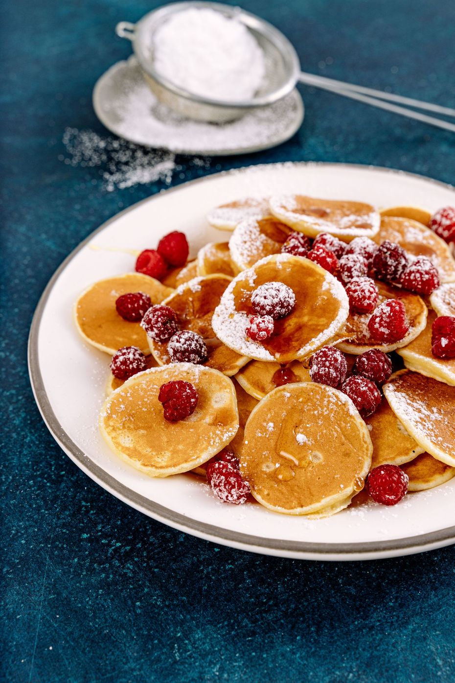 Plate of pancakes topped with berries and powdered sugar on a blue tablecloth, with a spoon and sieve nearby