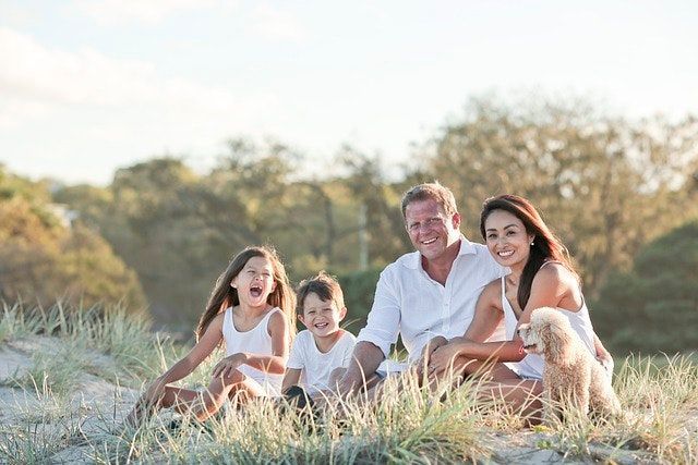 Family sits on the sand, smiling. They wear white, with a dog. Outdoors, near tall grass.