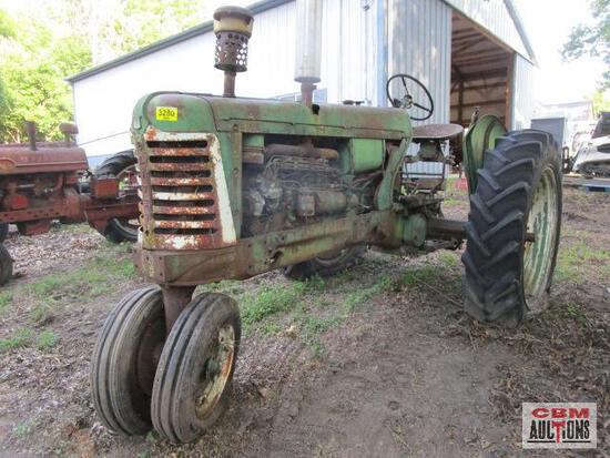 An old green tractor is parked in the dirt in front of a barn.