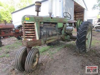 An old green tractor is parked in the dirt in front of a barn.