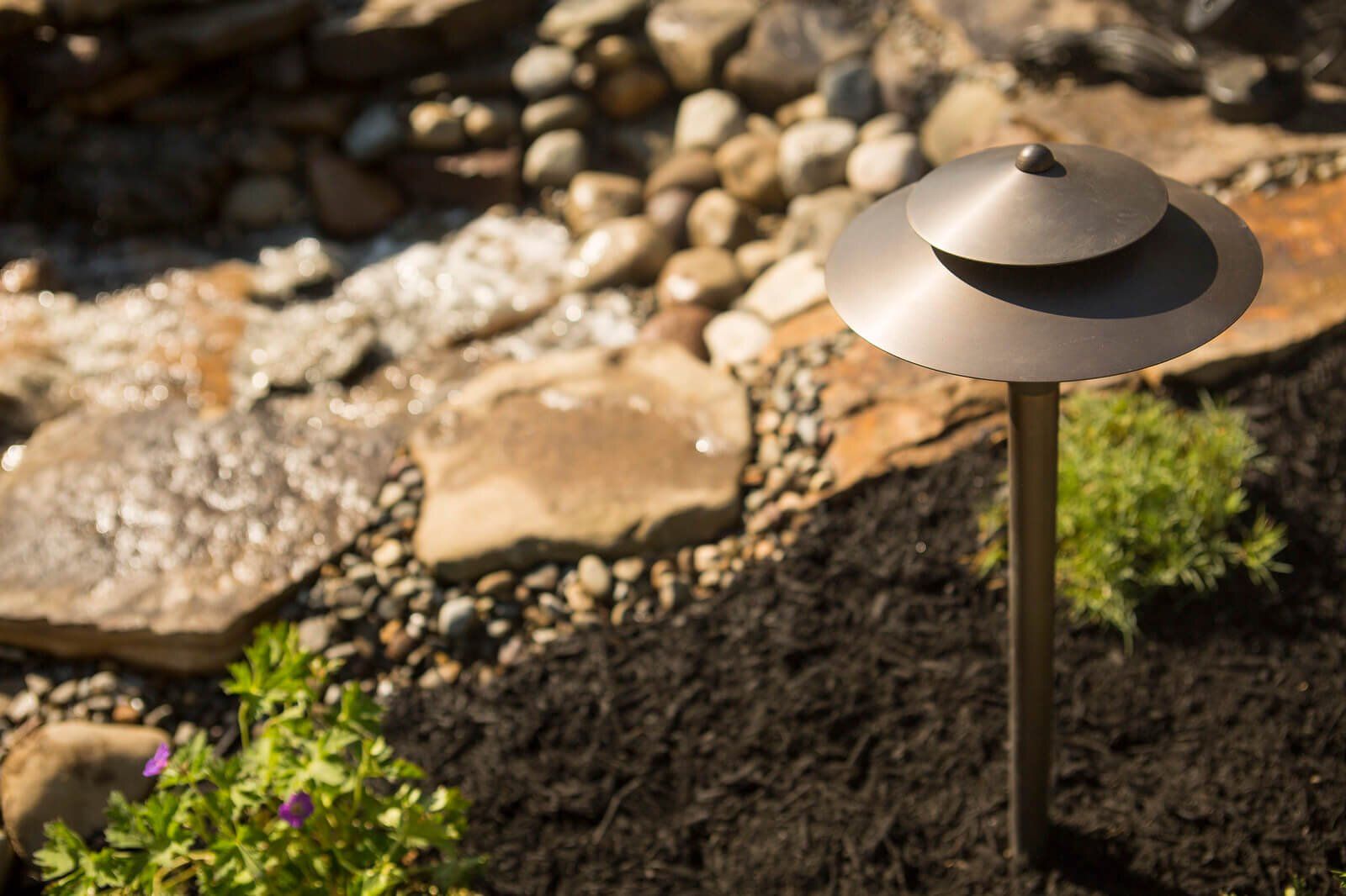 Garden path light beside rocks and small green plants in sunlight