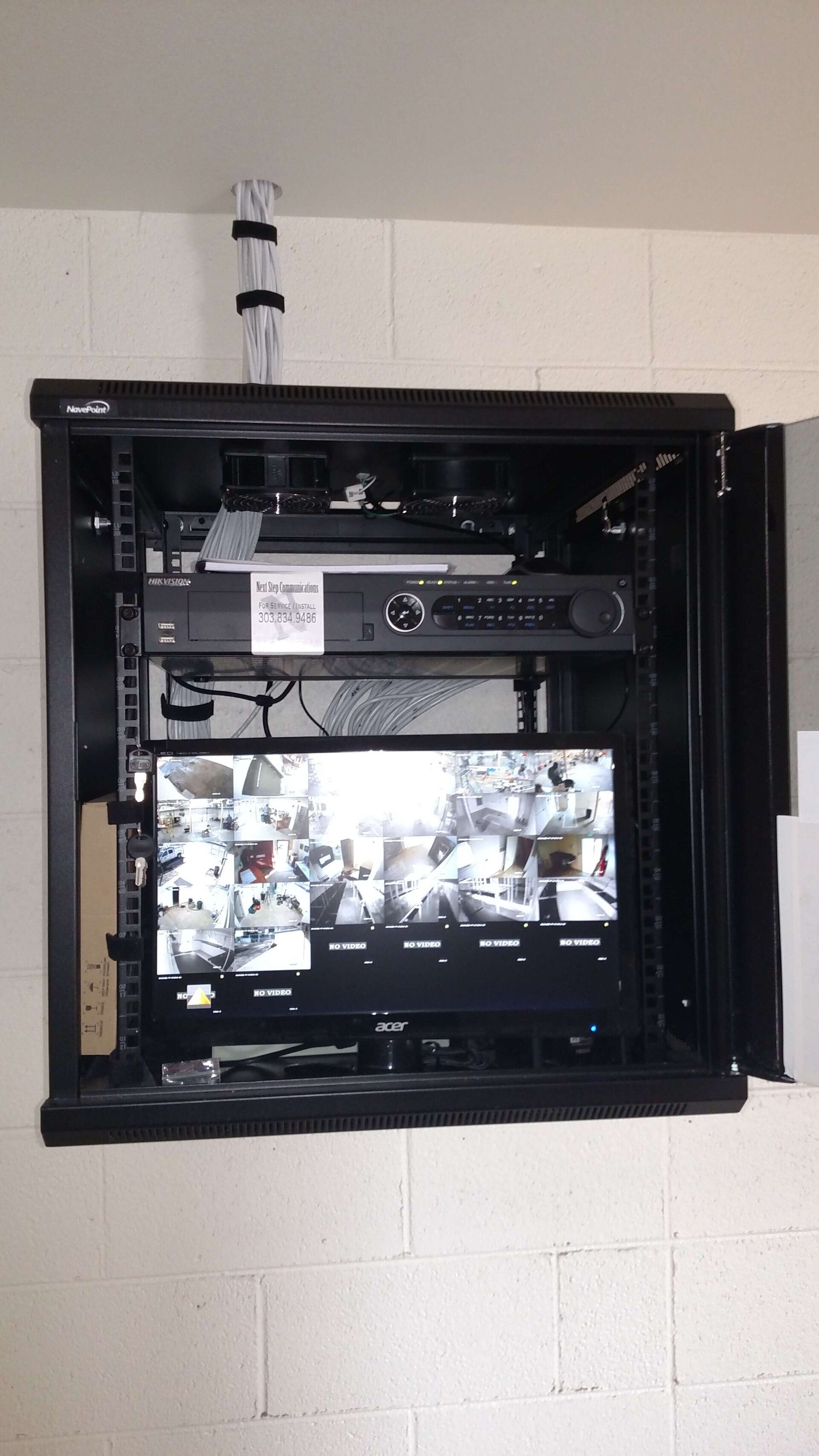 Open black equipment cabinet with electronics and a monitor inside on a white cinderblock wall.