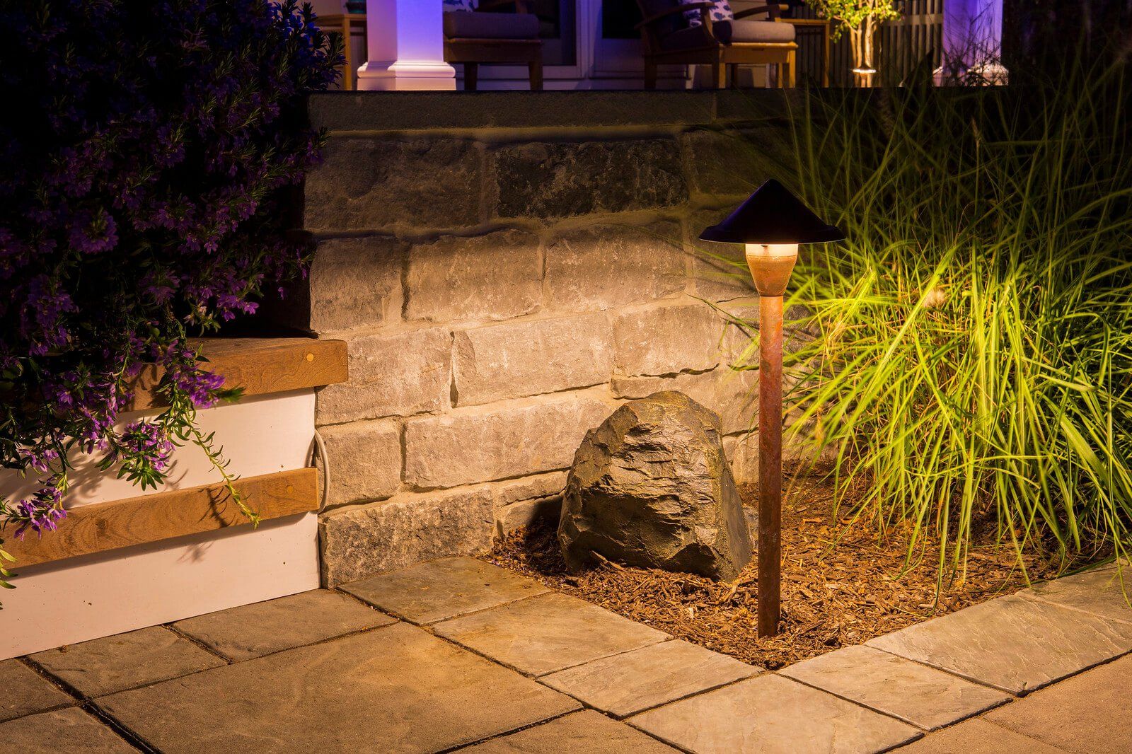 Garden path lit by a small lamp beside a stone wall and ornamental grass at night