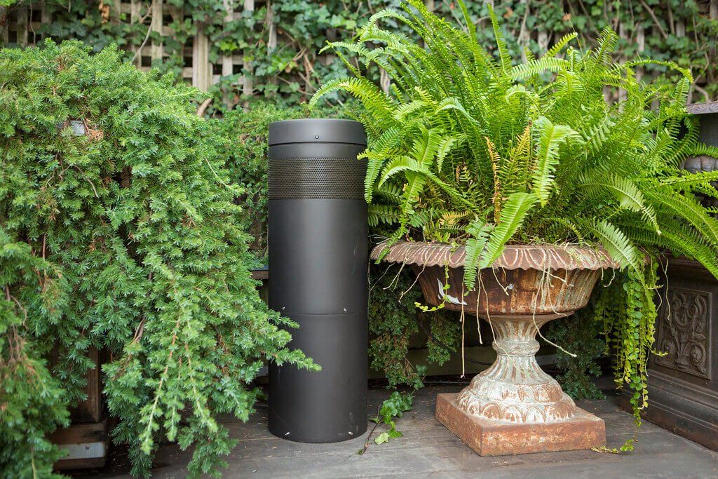 Black cylindrical bin beside a stone urn planter with lush green ferns in a garden setting