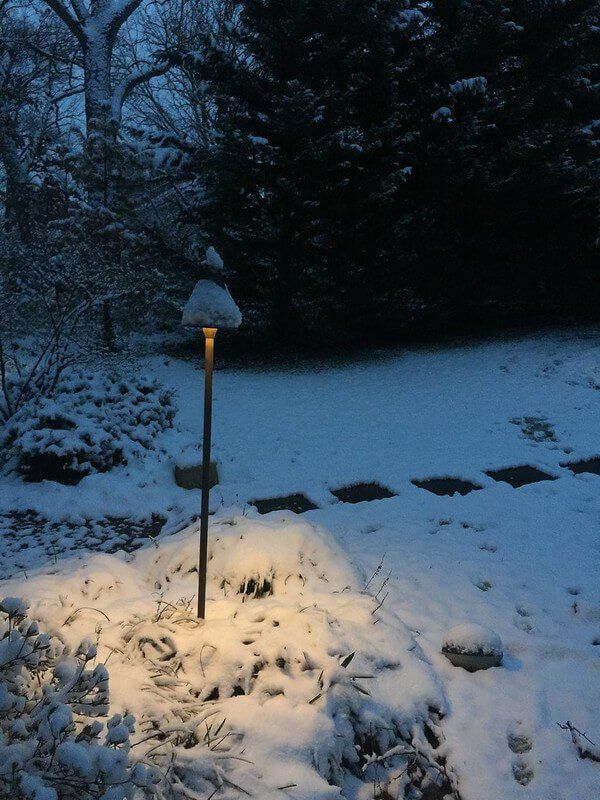 Snow-covered garden at dusk with a lit garden lamp casting warm light on the snowy ground.