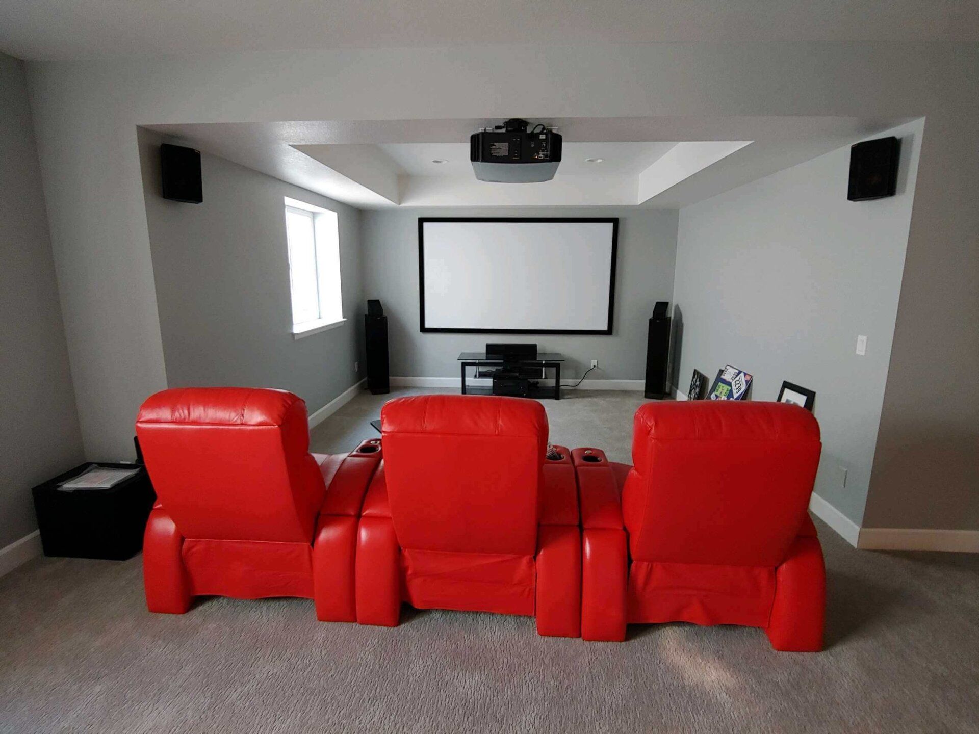 Home theater with three red recliners facing a projector screen and speakers in a bright room