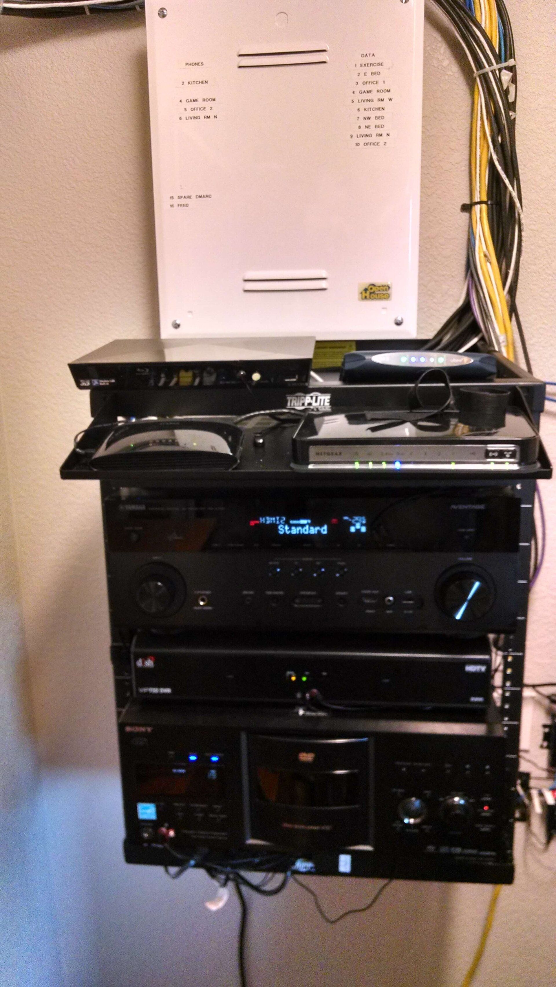 Stacked home audio equipment and cables mounted on a wall shelf, with a white utility box above.