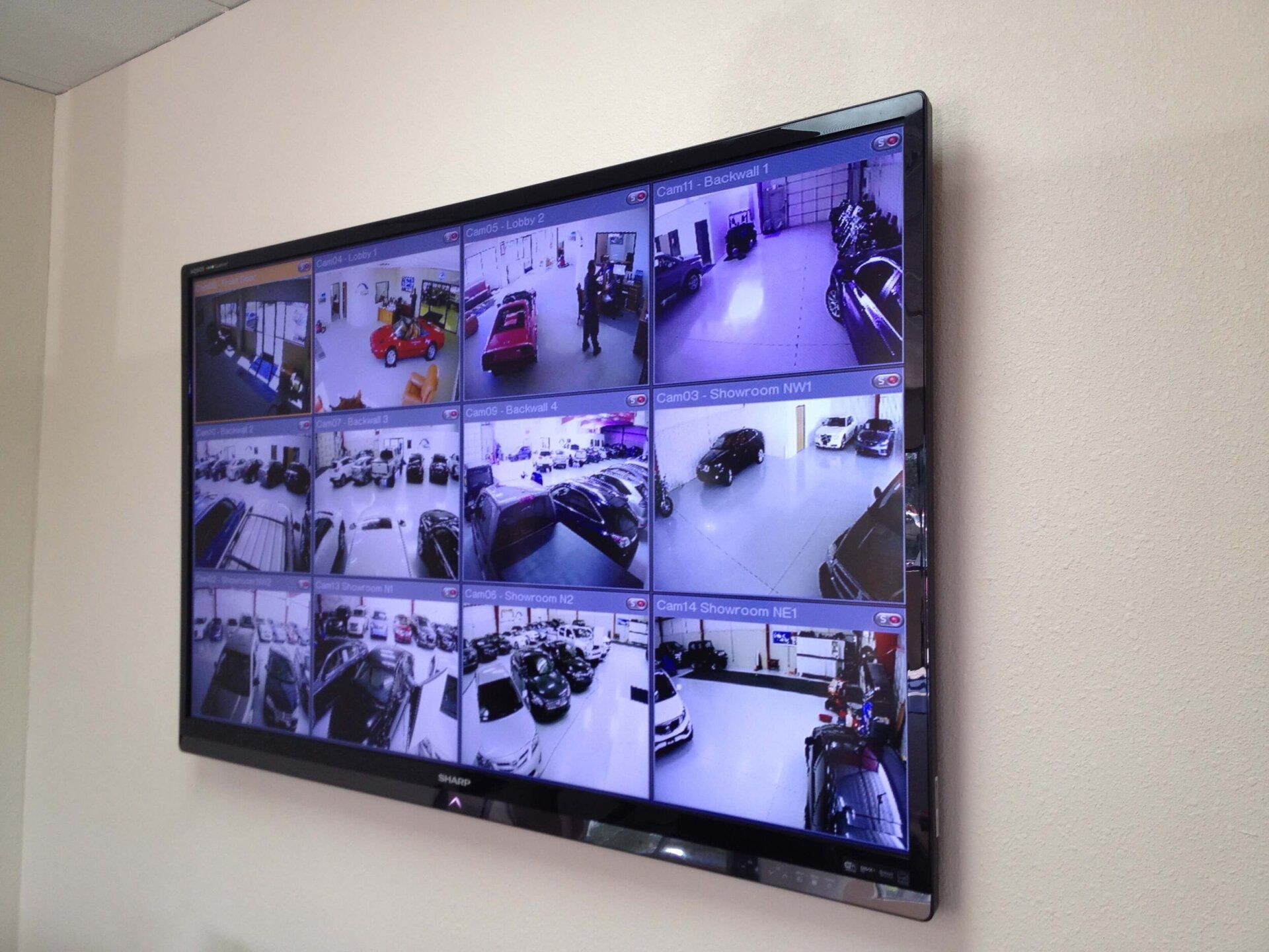 Wall-mounted monitor showing multiple CCTV camera feeds of a parking lot and building entrances