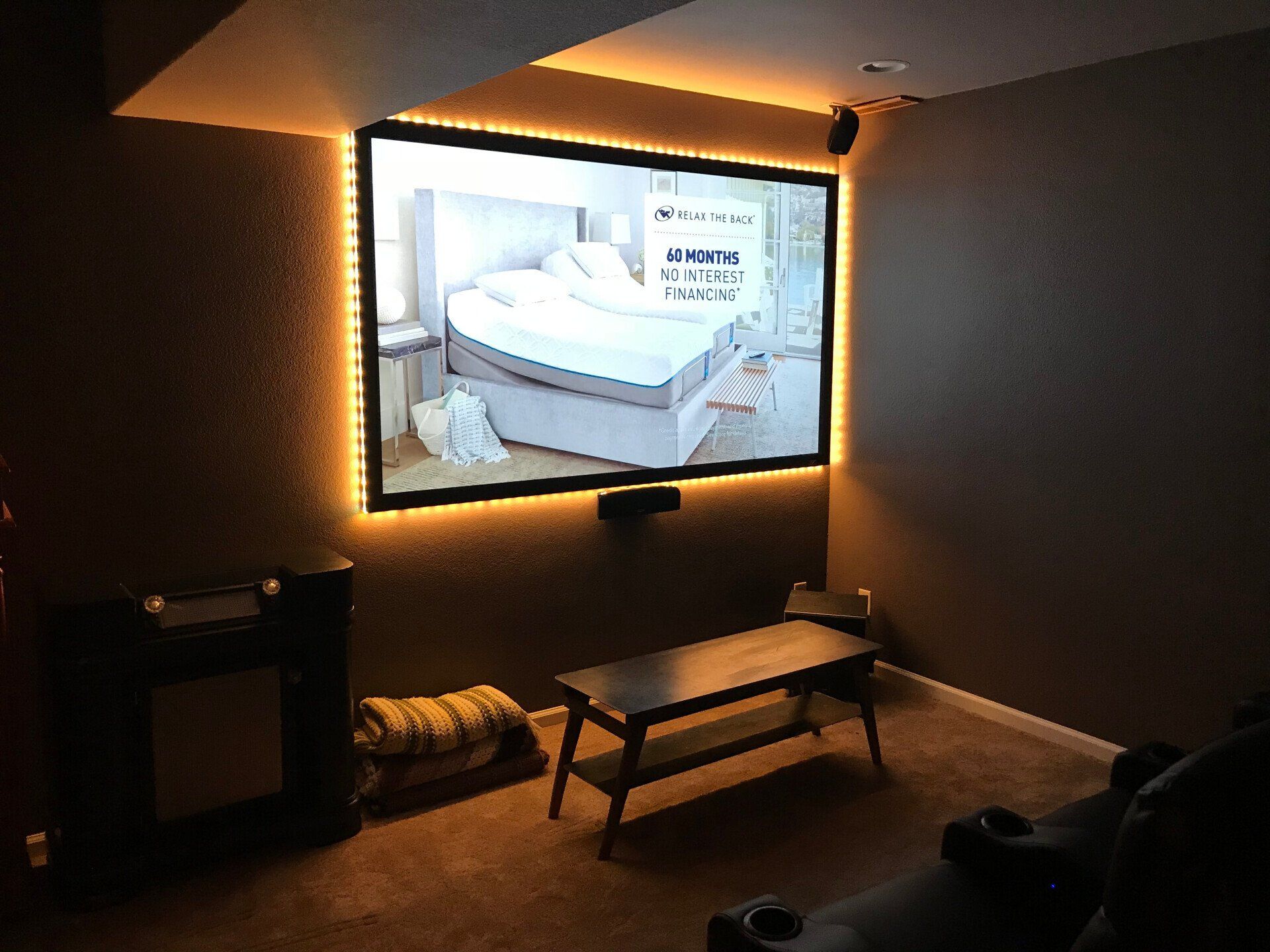 Dim room with a large wall-mounted TV showing a bright bedroom scene and a small bench in front.