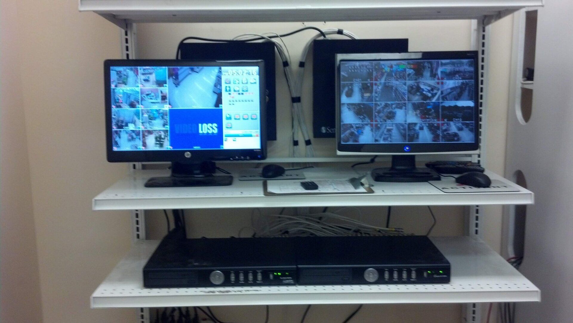 Surveillance monitor station with two screens showing camera feeds on white shelves with a black DVR below