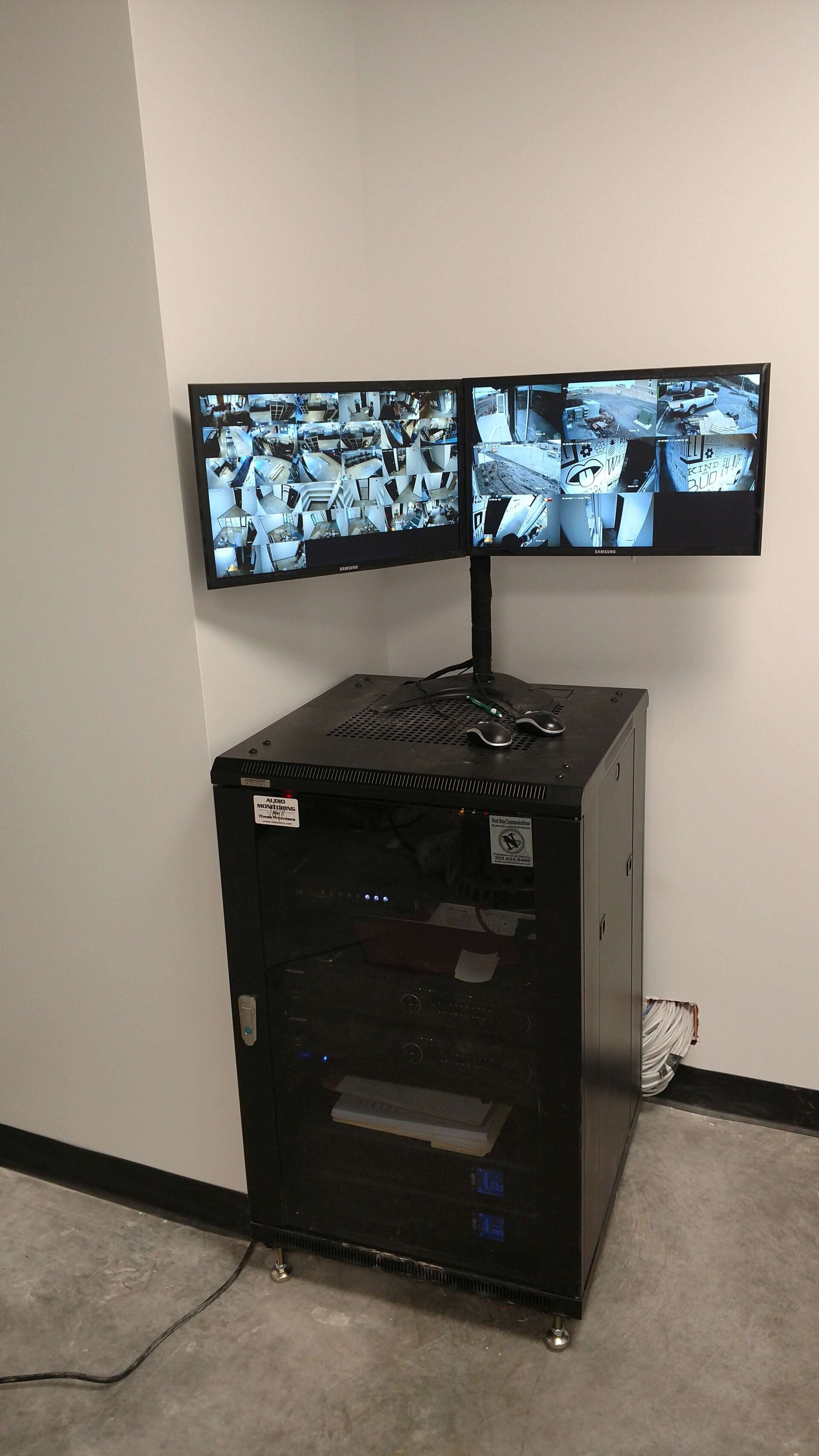 Black security cabinet with two monitors showing multiple CCTV camera feeds in a hallway.