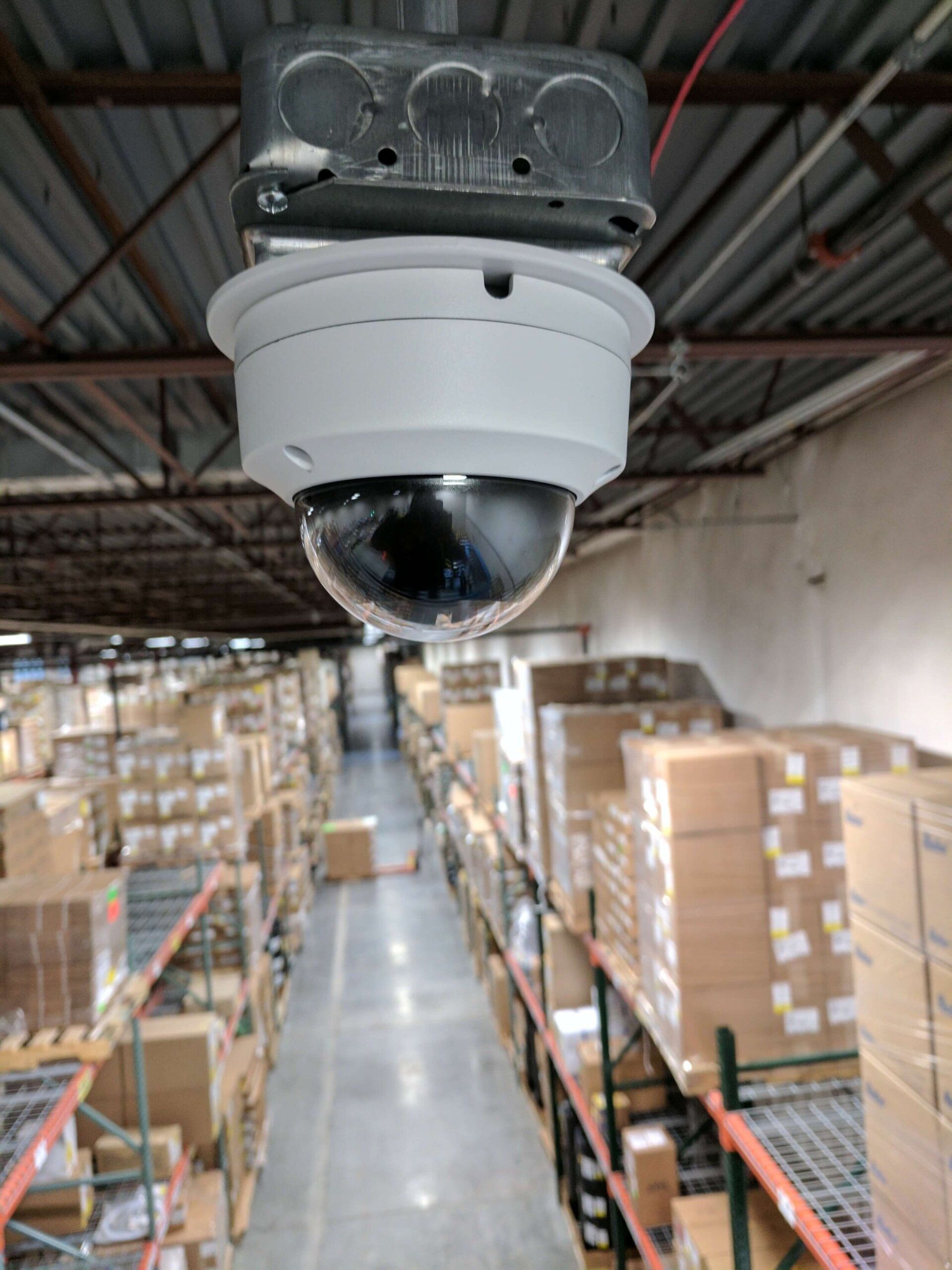Ceiling-mounted security camera overlooking a warehouse aisle with stacked boxes and shelving