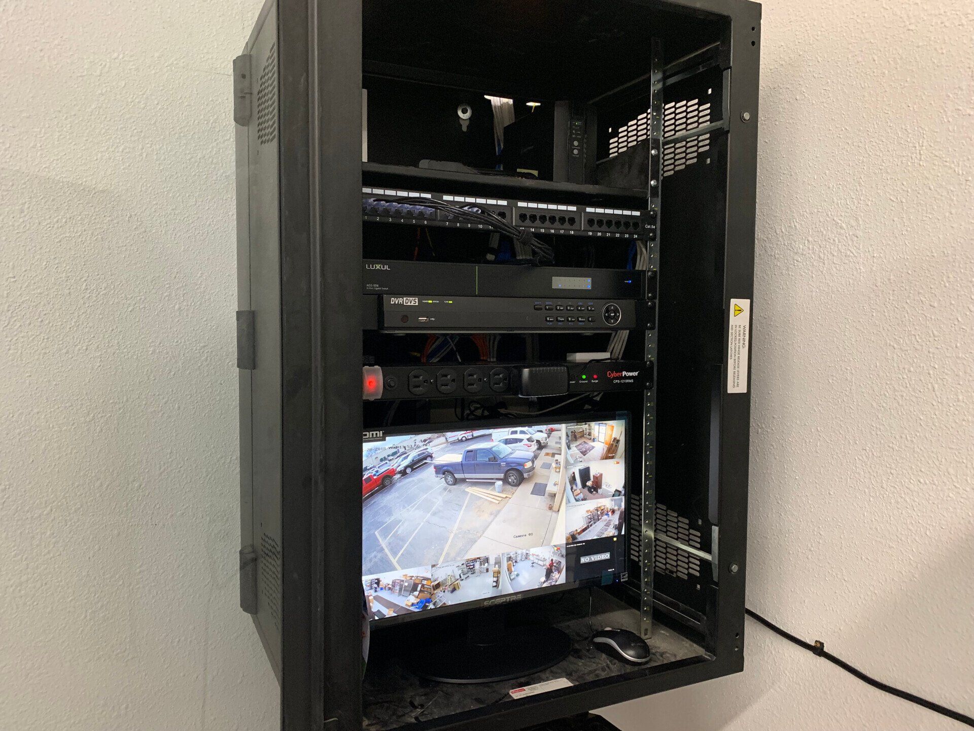 Black server rack with equipment and a monitor showing a surveillance camera view of a parking lot.
