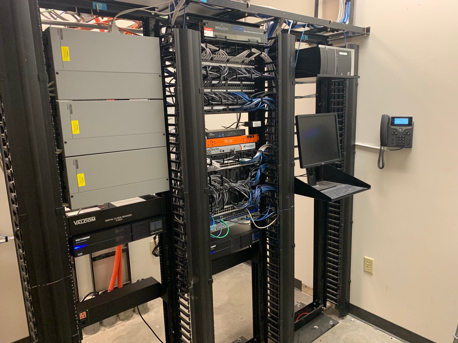 Server rack with network equipment, cables, and wall-mounted control panel in a data room.
