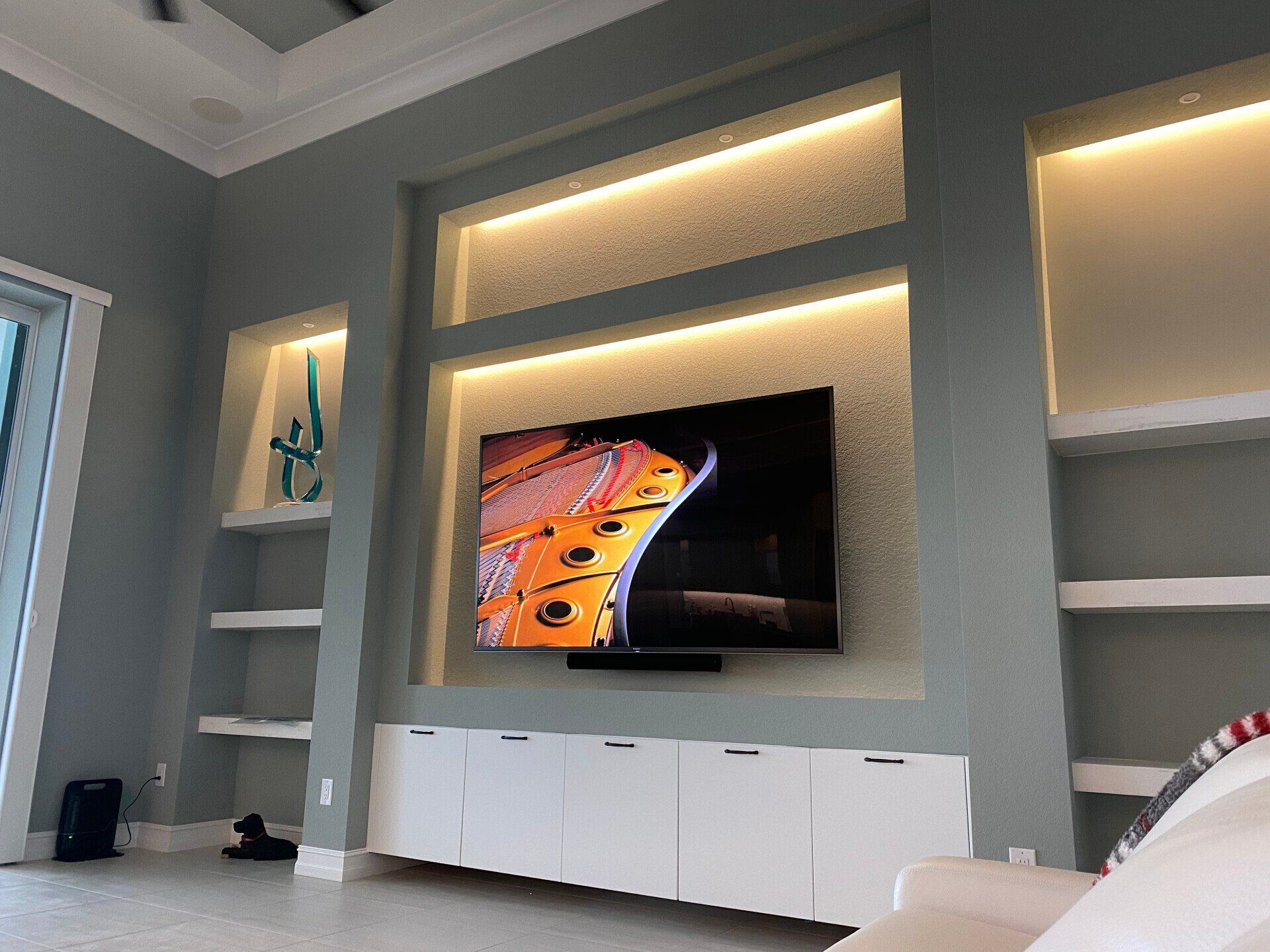Modern living room with wall-mounted TV, backlit shelves, and white cabinets under a gray wall
