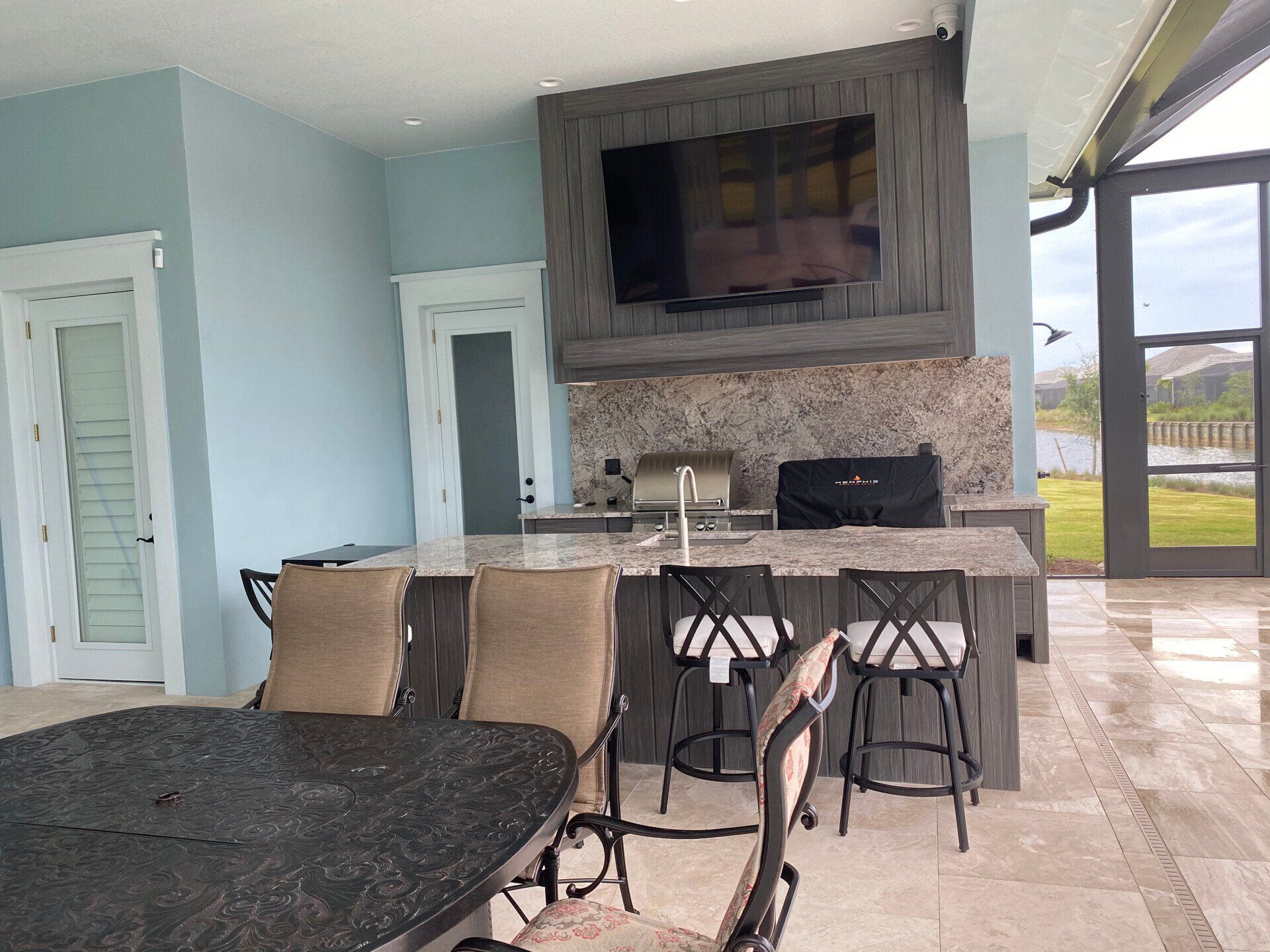 Patio seating area with table, bar stools, stone fireplace, and wall-mounted TV by the pool.