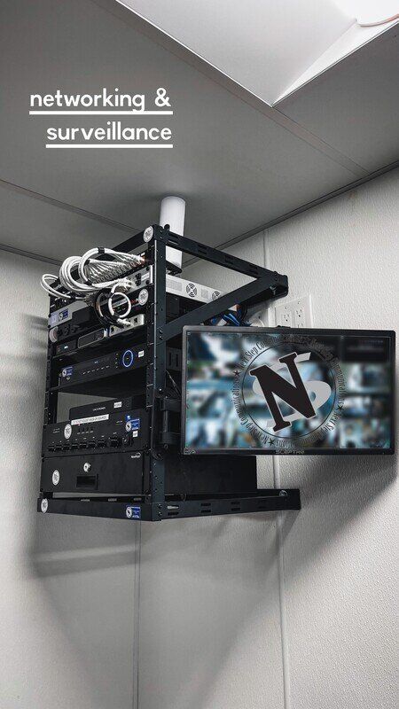 Wall-mounted networking and surveillance rack with cables and a monitor in a gray corner room