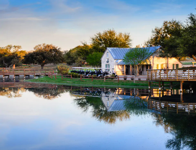 A white lakeside house with a porch and trees reflected in calm water at sunset