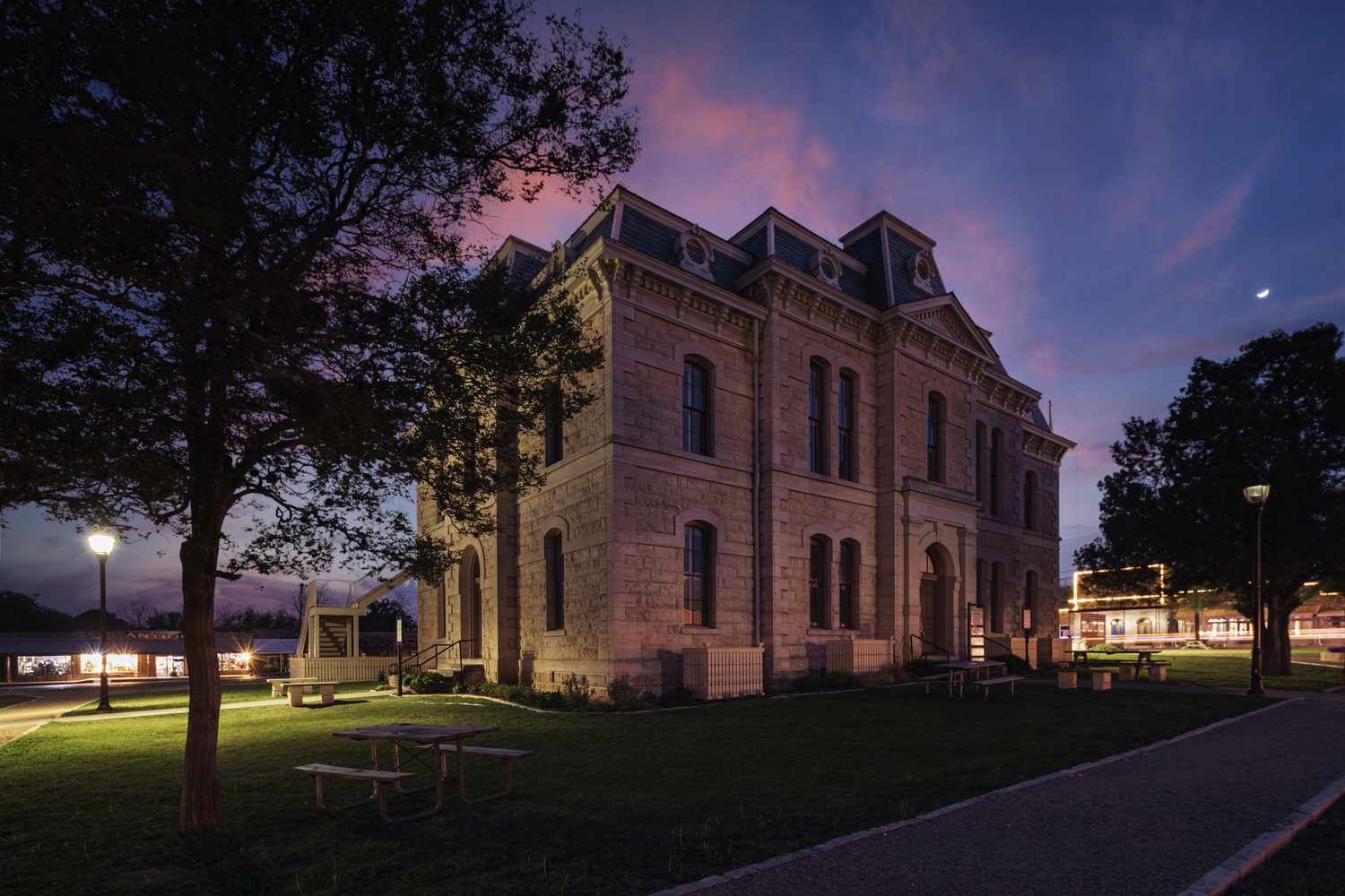 Stone building lit at dusk in a park, with trees and a path under a purple evening sky