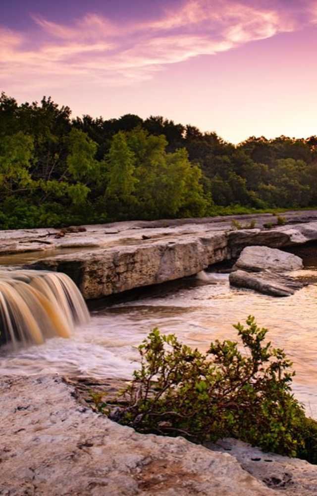 Waterfall over rocky ledge at sunset, with green trees and a pink sky.