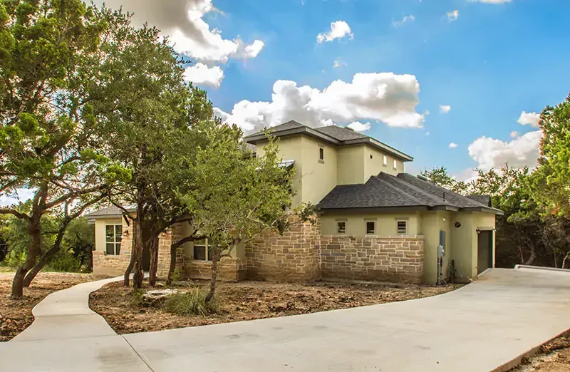 Large beige house with stone accents, curved driveway, and trees under a blue sky