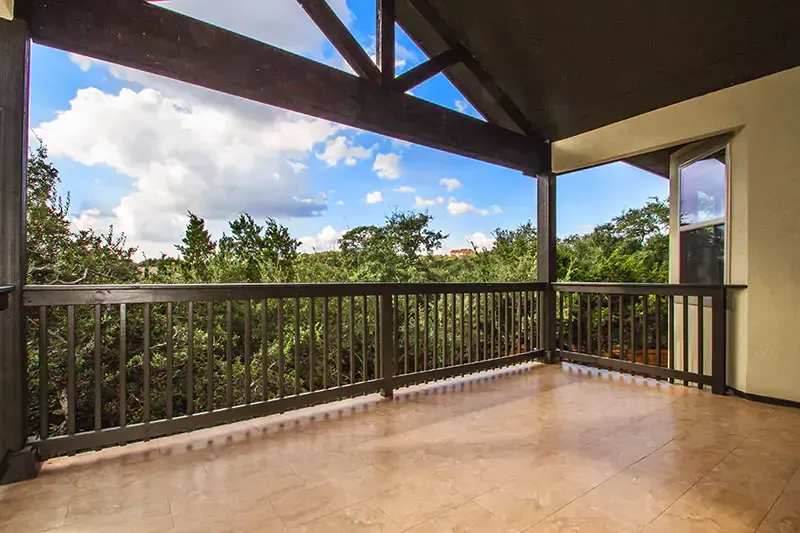 Covered balcony with wooden railing, tiled floor, and open view of trees and cloudy sky
