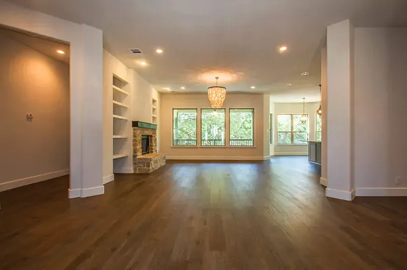 Empty living room with hardwood floors, recessed lights, white walls, and large windows with greenery outside