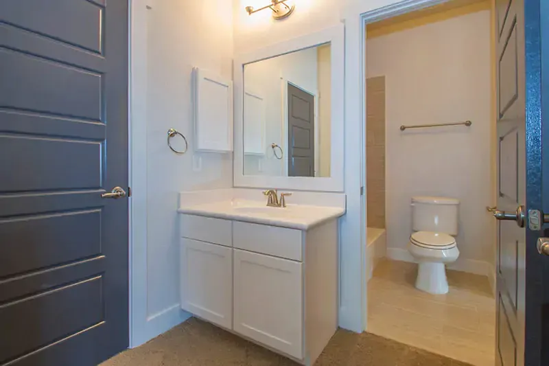 Small bathroom with white vanity, mirror, and toilet; open doorways on both sides.