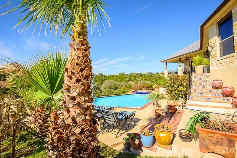 Sunlit backyard patio with palm trees, lounge chairs, and a blue swimming pool beside a stone house