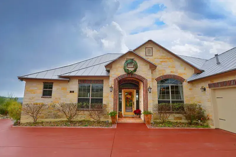 Single-story tan house with stone accents, red driveway, and a covered front entry under a cloudy sky