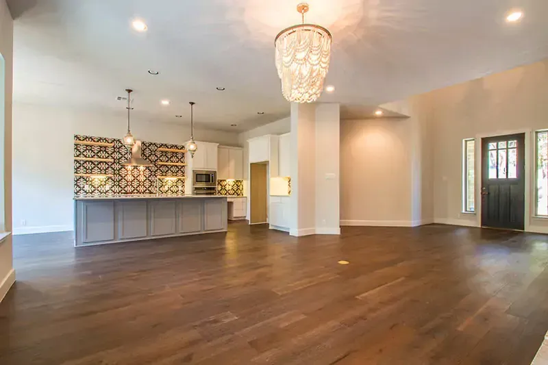 Empty open-plan living room with dark wood floors, white walls, kitchen island, and chandelier lighting