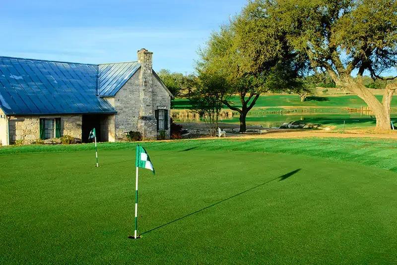 Golf green with flagstick beside a stone clubhouse and trees under a clear blue sky