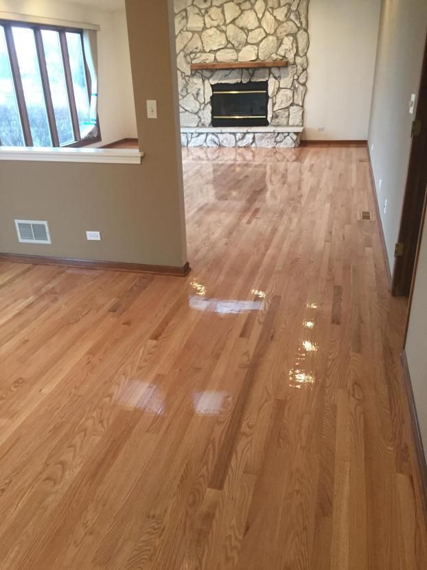 Hardwood floors in a living room, reflecting light. A stone fireplace and windows are in the background.