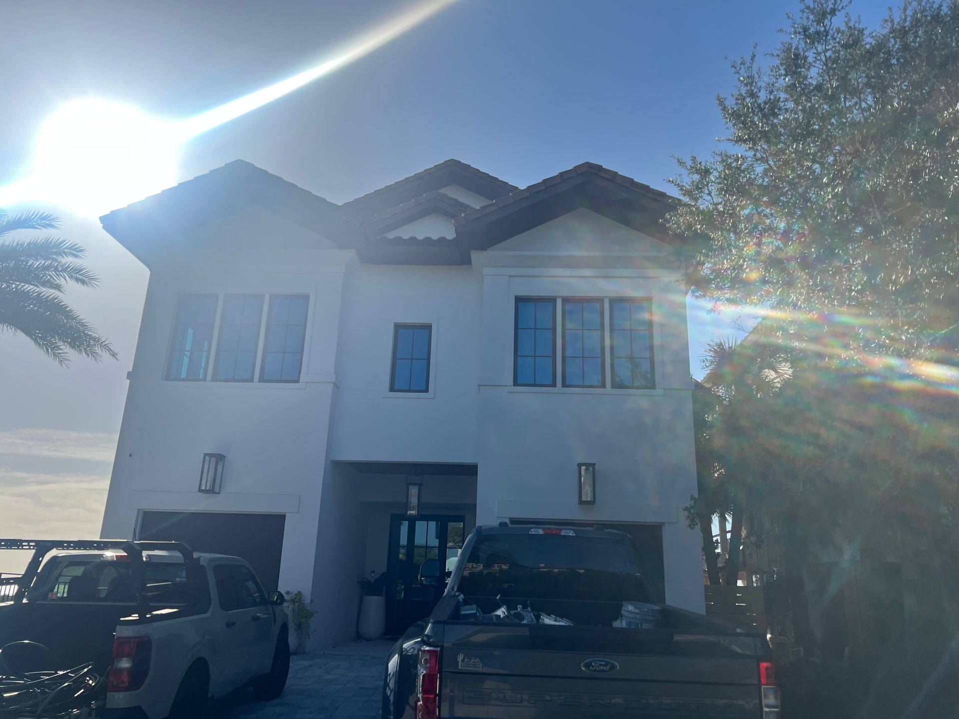 White two-story house with dark brown roof. Two trucks parked in front, bright sunlight.