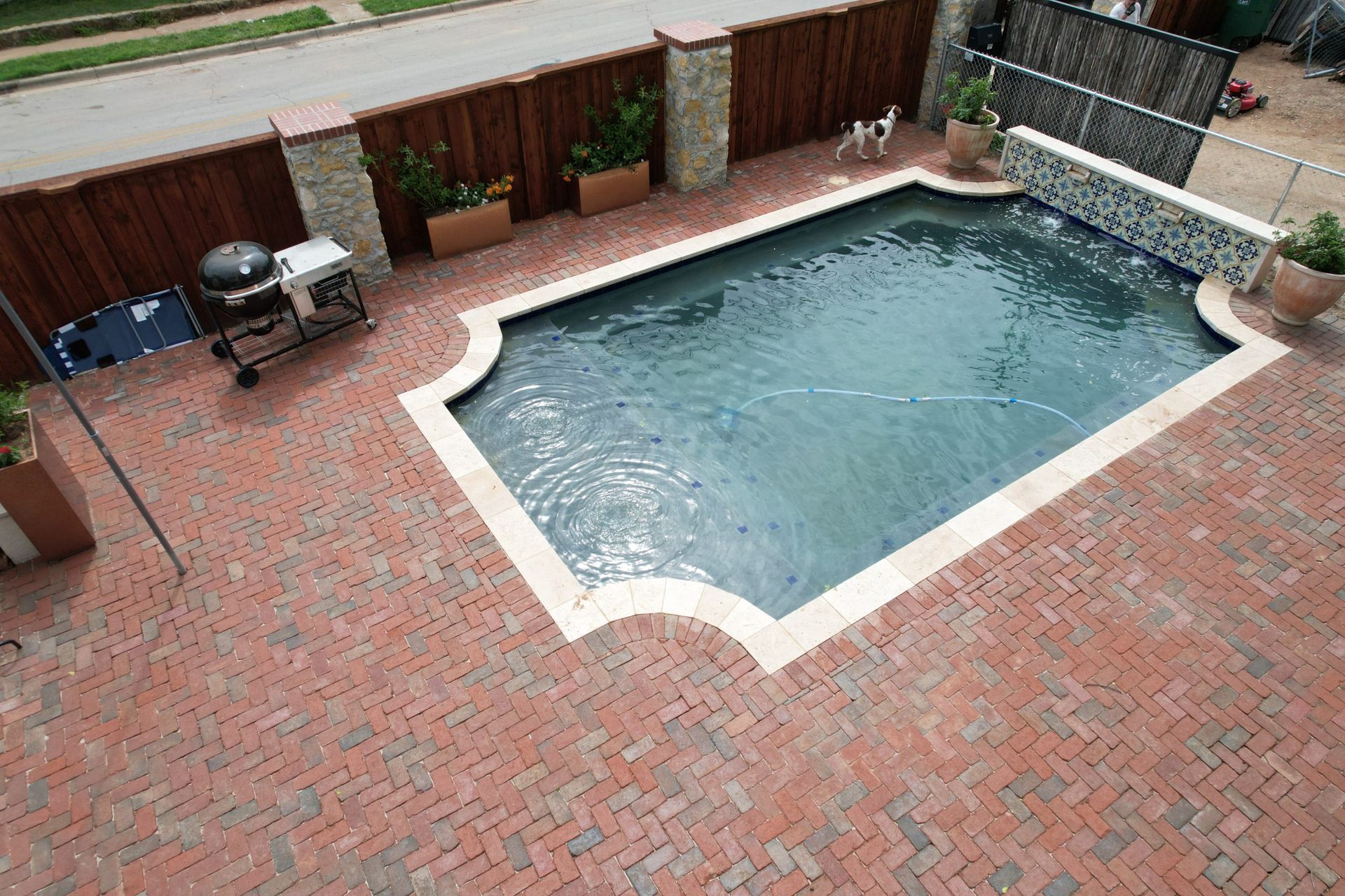Patio with a kidney-shaped swimming pool, brick paving, and wooden fence with potted plants