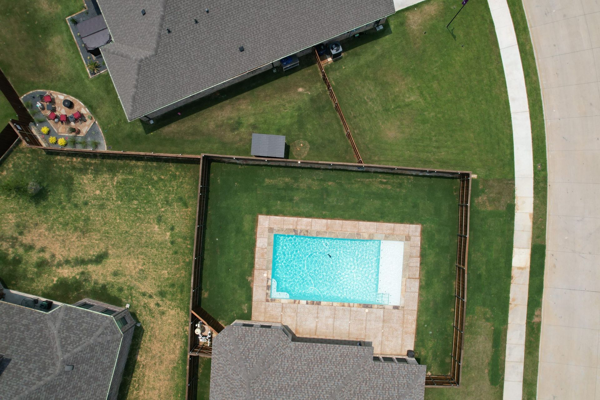 Aerial view of a backyard with a rectangular swimming pool inside a fenced yard near houses and a curved road.