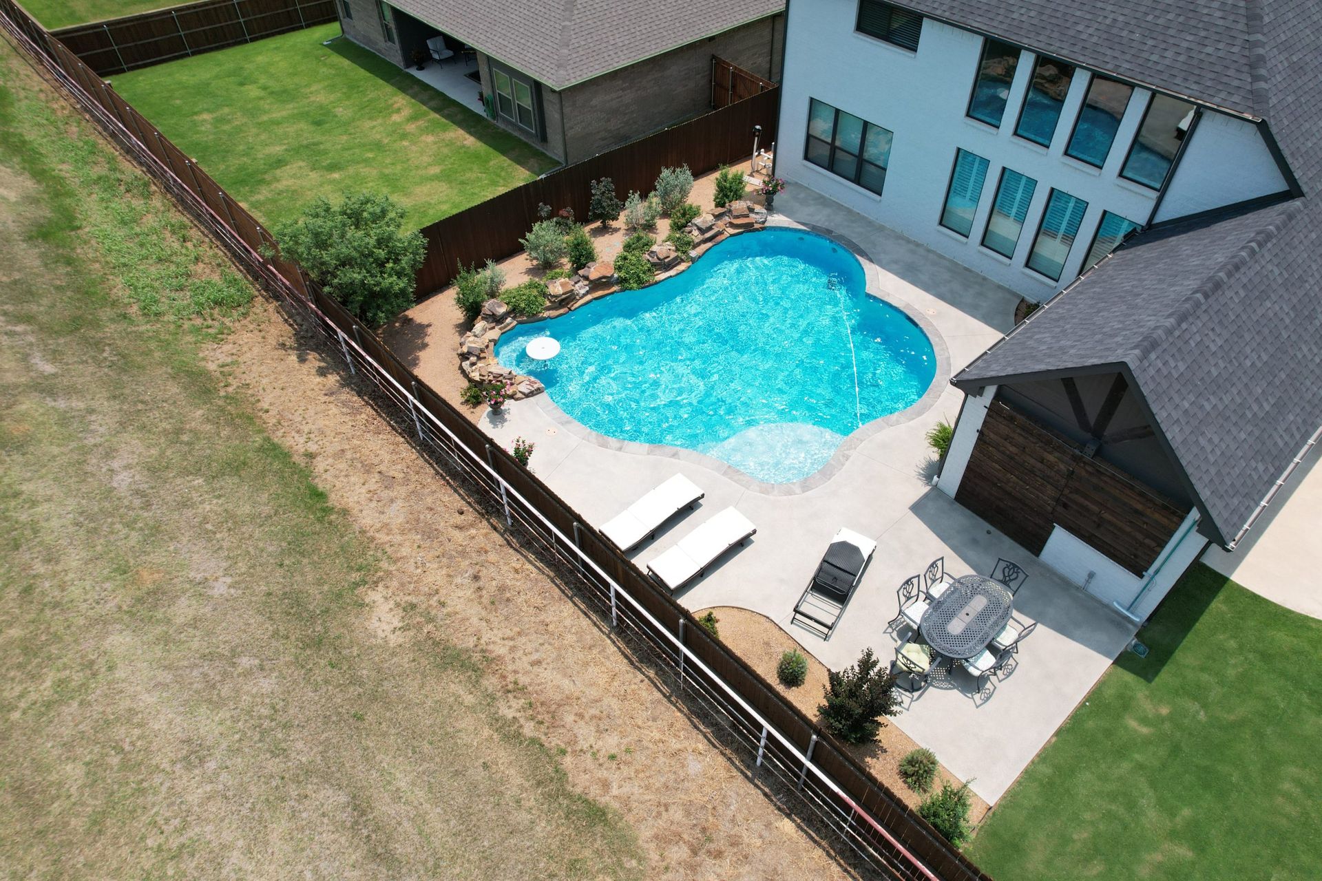 Aerial view of a backyard pool, patio chairs, and a house beside a fenced lawn.