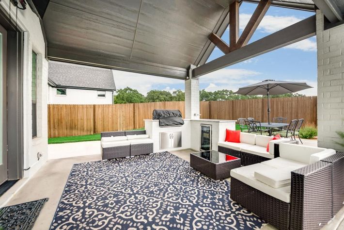 Covered backyard patio with wicker seating, patterned rug, and wooden fence.