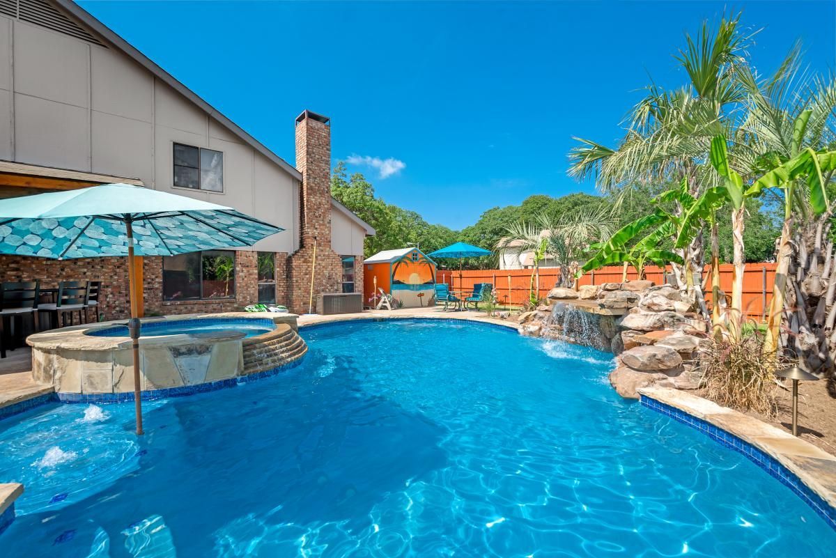 Backyard pool with blue water, stone spa, palm trees, and patio umbrellas under a sunny sky