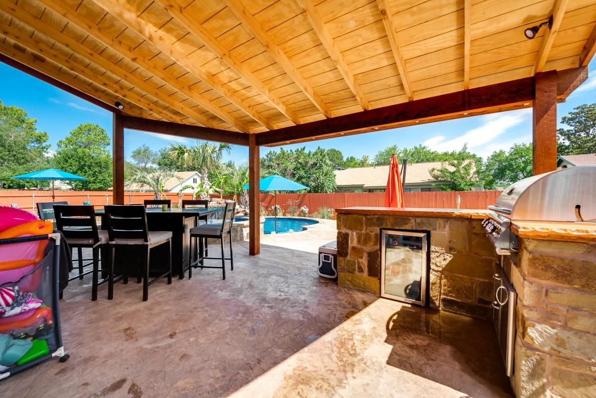 Covered patio with dining table, grill, and poolside seating under a wooden roof.