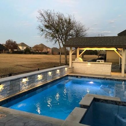 Backyard pool and spa at dusk with blue water and a lit pavilion beside a fence and leafless tree