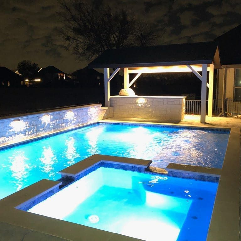 Illuminated backyard pool and hot tub at night with a lit gazebo and stone wall fountain