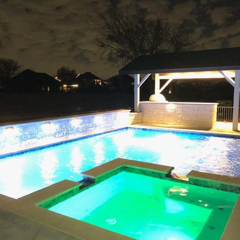 Nighttime backyard pool with a glowing green hot tub and lit swimming pool under a covered patio.