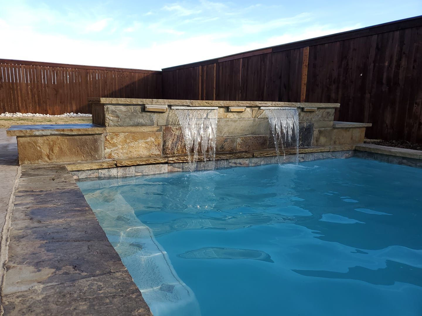 Backyard pool with a stone waterfall and wooden fence under a blue sky