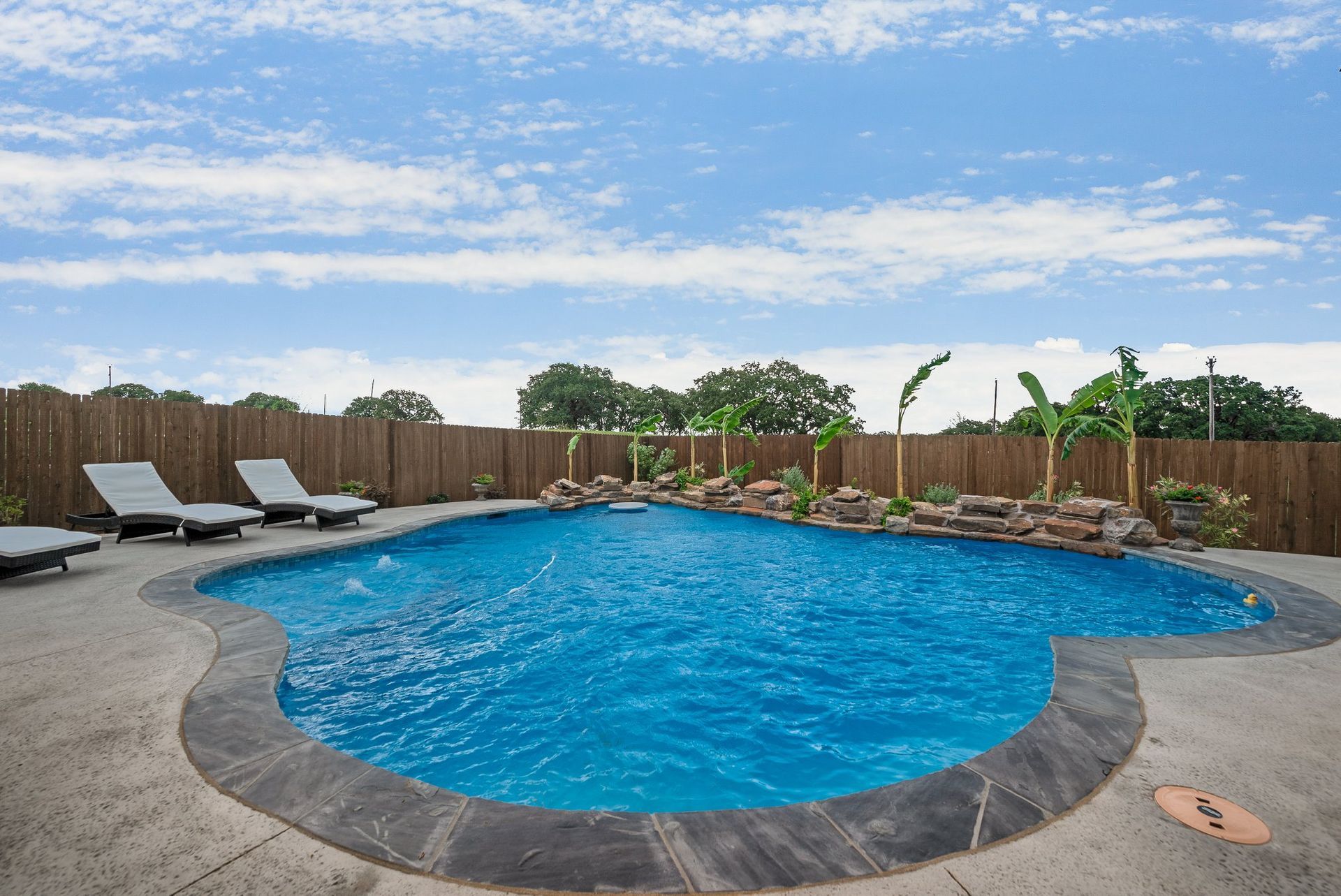 Backyard swimming pool with blue water, lounge chairs, and a wooden fence under a cloudy sky