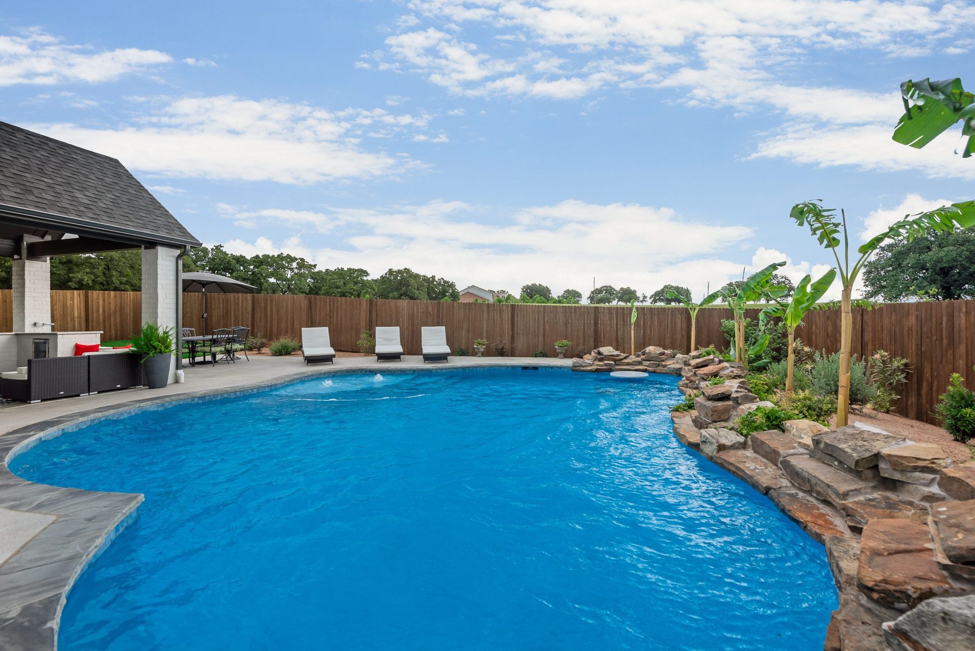 Backyard swimming pool with a stone waterfall, lounge chairs, and a covered patio under a blue sky