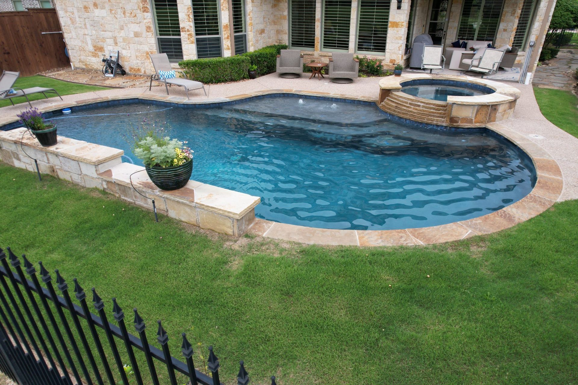Backyard swimming pool with stone patio, hot tub, and green lawn behind a black fence